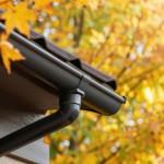 House corner with dark gutter and downspout under a tiled roof, autumn leaves in the background.